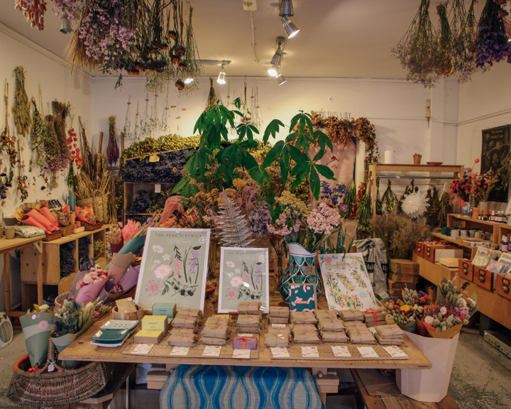 Interior of a boutique shop filled with dried flowers, plants, and botanical arrangements. A central wooden table displays illustrated floral prints, seed packets, and small items, while shelves and walls around the room are decorated with hanging bunches of dried flowers and greenery. The space has warm lighting and a natural, rustic aesthetic.
