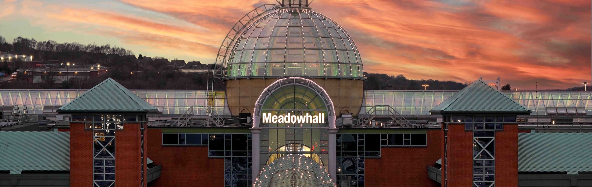 The Meadowhall exterior at sunset, featuring the glass-domed main entrance, red-brick walls and a dramatic orange and pink sky.