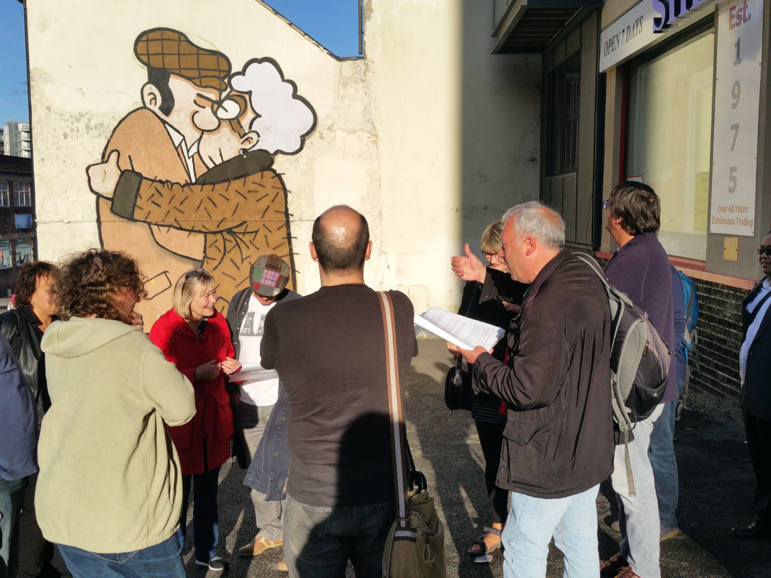 Crowd of people stood looking at 'The Snog' by Pete McKee on the side of Fagan's pub