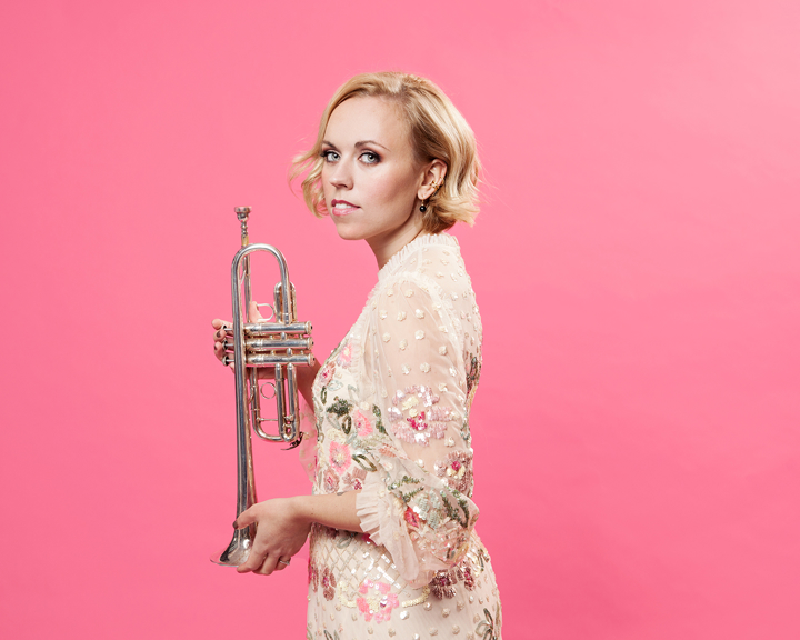 A woman holding a trumpet poses against a bright pink wall.