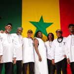 A group of nine musicians pose together in front of a large Senegal flag featuring green, yellow and red vertical stripes with a green star at the centre. Most wear matching white tunics with black trim, while one person in the middle wears a sleeveless white outfit. The group stands closely together, smiling and facing the camera, creating a warm and unified portrait.