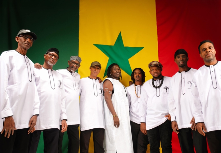 A group of nine musicians pose together in front of a large Senegal flag featuring green, yellow and red vertical stripes with a green star at the centre. Most wear matching white tunics with black trim, while one person in the middle wears a sleeveless white outfit. The group stands closely together, smiling and facing the camera, creating a warm and unified portrait.