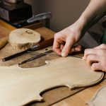 Close-up of hands shaping thin strips of wood on a violin body at a woodworking bench. The unfinished instrument rests among specialist tools, including chisels, a knife and a carving gouge. A wood slice and workshop equipment sit in the background. Warm lighting highlights the craftsmanship and detailed process of traditional stringed instrument making.
