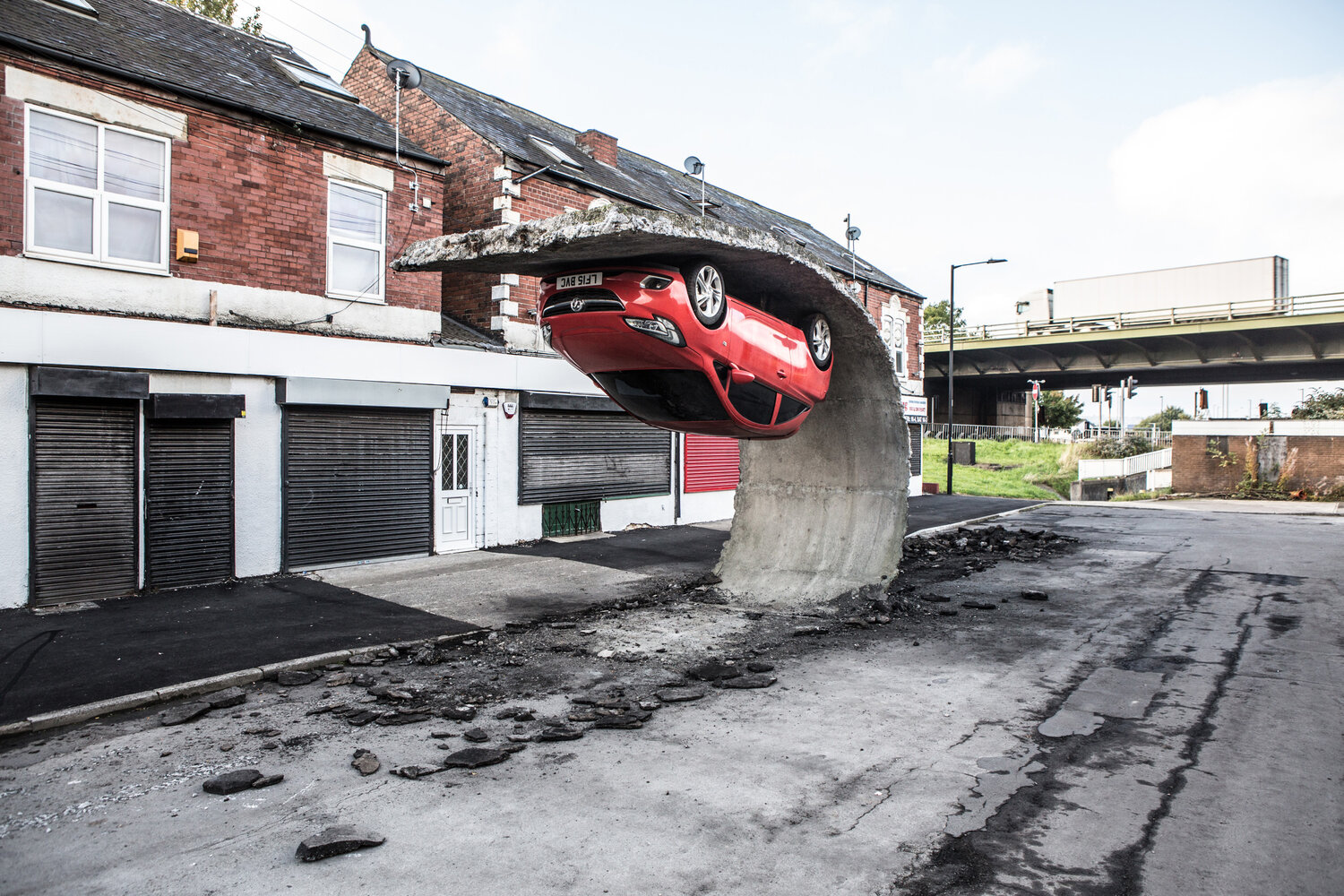 Red car embedded upside down in a large curved concrete structure on a deserted street, with terraced brick buildings and closed shopfronts in the background.